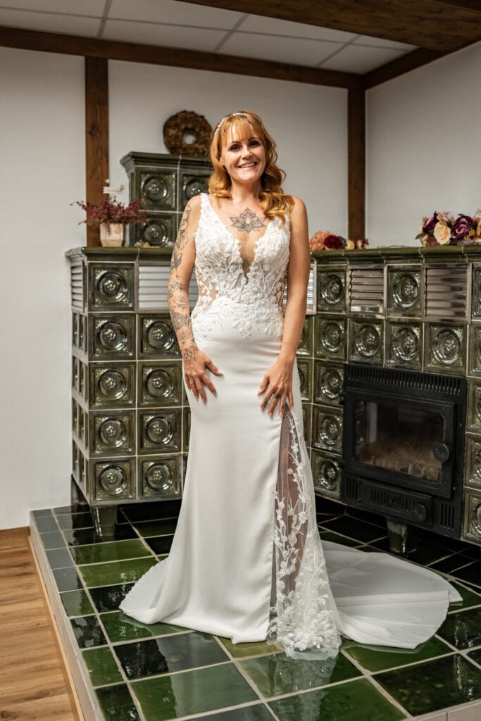 Lächelnde Frau im weißen Brautkleid vor Kachelofen.