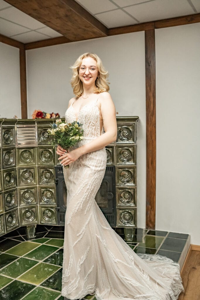Frau im Brautkleid mit Blumenstrauß lächelt vor Ofen.