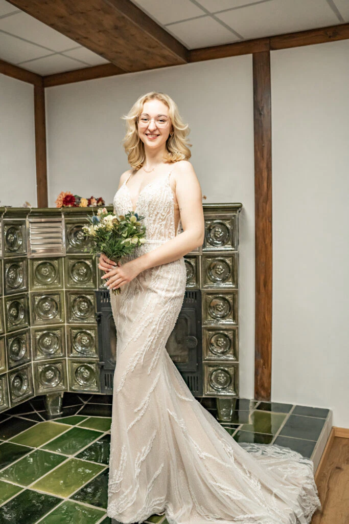 Frau im Brautkleid mit Blumenstrauß lächelt vor Ofen.
