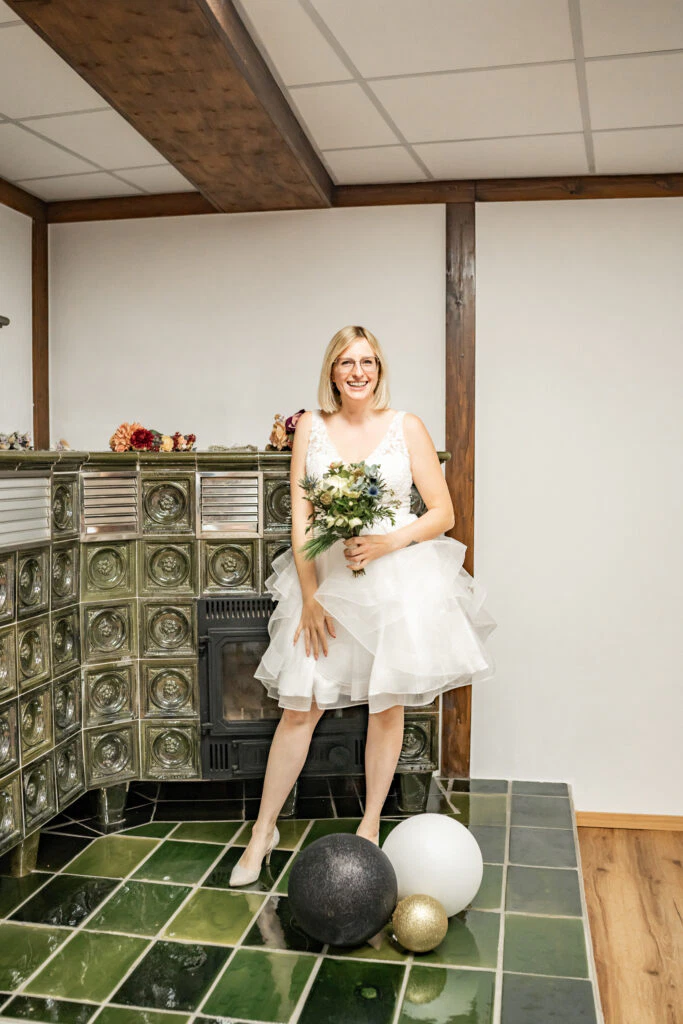 Braut mit Blumenstrauß vor Kachelofen lächelt